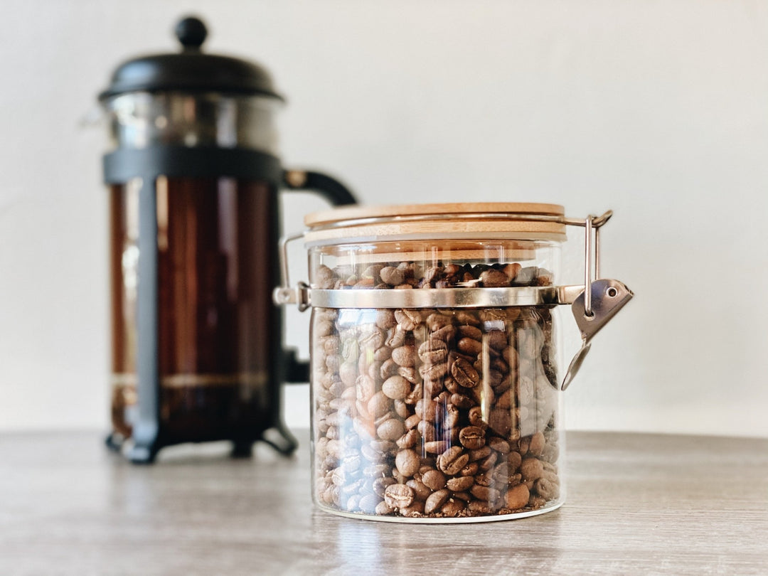 glass jar with coffee beans and coffee plunger in the background