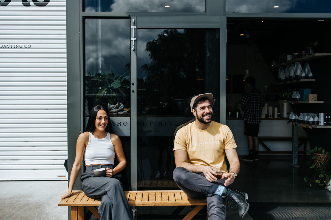 woman and man sitting on a bench in front of a coffee roastery holding a cup of coffee in their hands